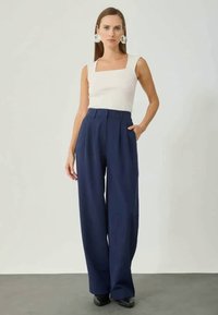 Witte top met vierkante hals, gecombineerd met hoog-gewijze marineblauwe wijde broek. Het model draagt grote zilveren oorbellen en zwarte schoenen. Glad textuur.