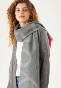 Grijze geweven sjaal met een patroonontwerp, voorzien van roze franjes aan de uiteinden, over een grijze cardigan gedrapeerd. Gladde textuur.