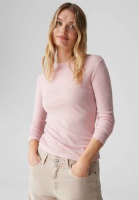 Hellrosa Langarmshirt mit rundem Ausschnitt, schlanker Passform und weicher Textur. Das Model hat die Hände in den Taschen und trägt hellbeige Hosen.