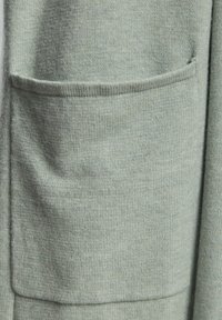 Vaaleanvihreä neulekangas, jossa on sileä pinta ja yksittäinen taskupatentti. Materiaali näyttää pehmeältä ja hieman ryppyiseltä.