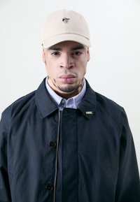 Light beige cap with logo, navy jacket featuring buttons and a zip, over blue and white striped shirt. Smooth fabric textures visible.