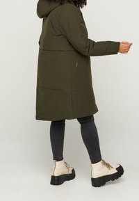 Długi oliwkowozielony płaszcz z kapturem, o gładkiej fakturze i bocznych kieszeniach na zamek. Noszony z ciemnymi dżinsami i kremowymi, masywnymi butami.