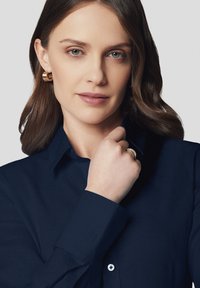 Granatowa koszula z kołnierzykiem, długimi rękawami, guzikami przy mankietach i subtelnym detalem z logo. Gładka faktura materiału. Złote kolczyki w kształcie kół.