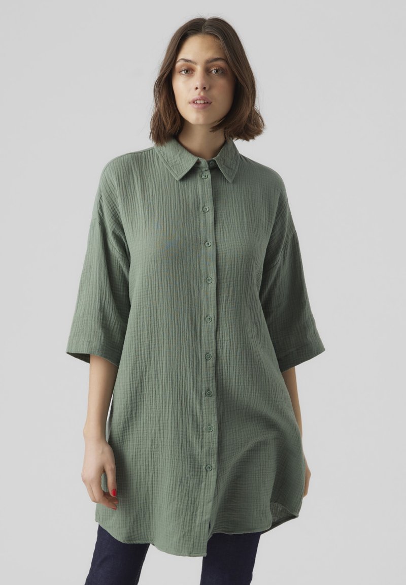 Chemise surdimensionnée verte à boutons, fabriquée dans un tissu texturé, dotée d'un col et de manches trois-quarts, portée sur un jean sombre.