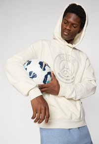 Sweat com capuz bege com gráfico "PARIS SAINT-GERMAIN", tecido texturizado e cordão. Segurando uma bola de futebol branca e azul com padrão.