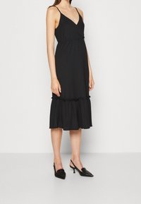 Robe noire midi avec un décolleté en V et des bretelles réglables, présentant un ourlet froncé et volanté. Texture lisse avec une coupe décontractée.