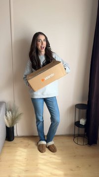 Karton doos met Zalando-logo, grijze trui, blauwe spijkerbroek en bruine slippers met witte bontafwerking. Binnensetting met lichtgekleurde muren.