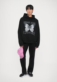 Melns pārāk liels kapuci, kas izceļ tauriņu grafiku un tekstu "Versace Jeans Couture", apvienots ar melnām biksēm un rozā cepuri.