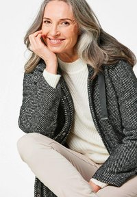 Zwart-wit herringbone-patroon jas met een textuurafwerking, gedragen over een crème kabelgebreide trui, gecombineerd met lichtbeige broek.