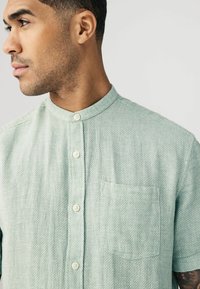 Camisa de manga corta en verde menta, con cuello mao, tejido texturizado y bolsillo frontal con un patrón sutil y botones blancos.