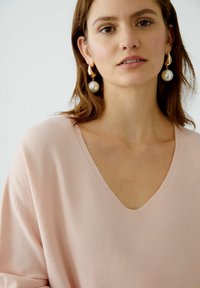 Light pink, waffle-textured knit top with a V-neckline. Model wears large pearl drop earrings and has medium-length hair.