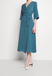 Femme portant une robe midi enveloppante teal avec ceinture à nouer et manches trois-quarts, associée à des escarpins blancs avec bride arrière.