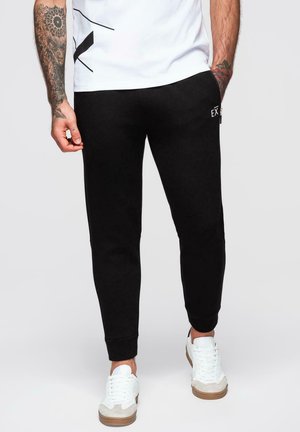 Svarta sweatpants gjorda av mjukt material, med tapered ben, elastiska muddar och en liten logotyp på höften. Bärs med vita sneakers.