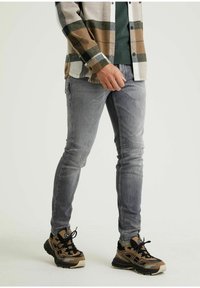 Bleka grå skinny jeans med lätt slitningar och dragkedjedetaljer, kombinerat med en mönstrad brun och grön rutig skjorta samt bruna sneakers.