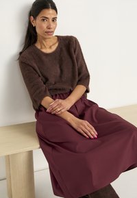Bruine pluchen trui met driekwartmouwen, gecombineerd met een bordeauxrode hoge taille rok. Subtiele stiksels op de rok; handen met gouden ring op een houten bank.