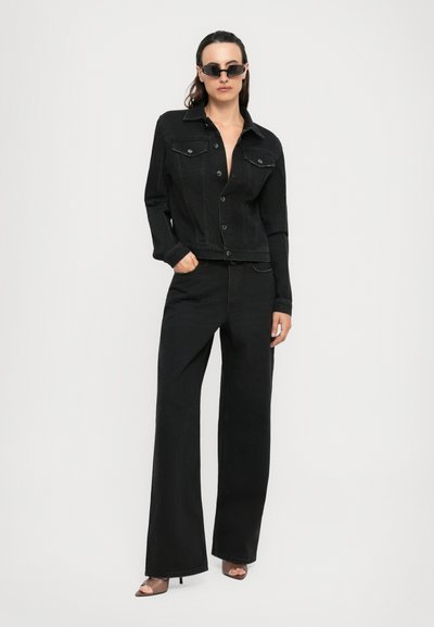 Woman wearing black denim jacket and wide-leg black jeans with brown open-toe heels and black sunglasses against white background.