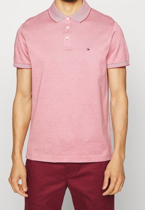 Homme portant un polo rose clair à manches courtes avec trois boutons et un petit logo, associé à un pantalon rouge foncé.