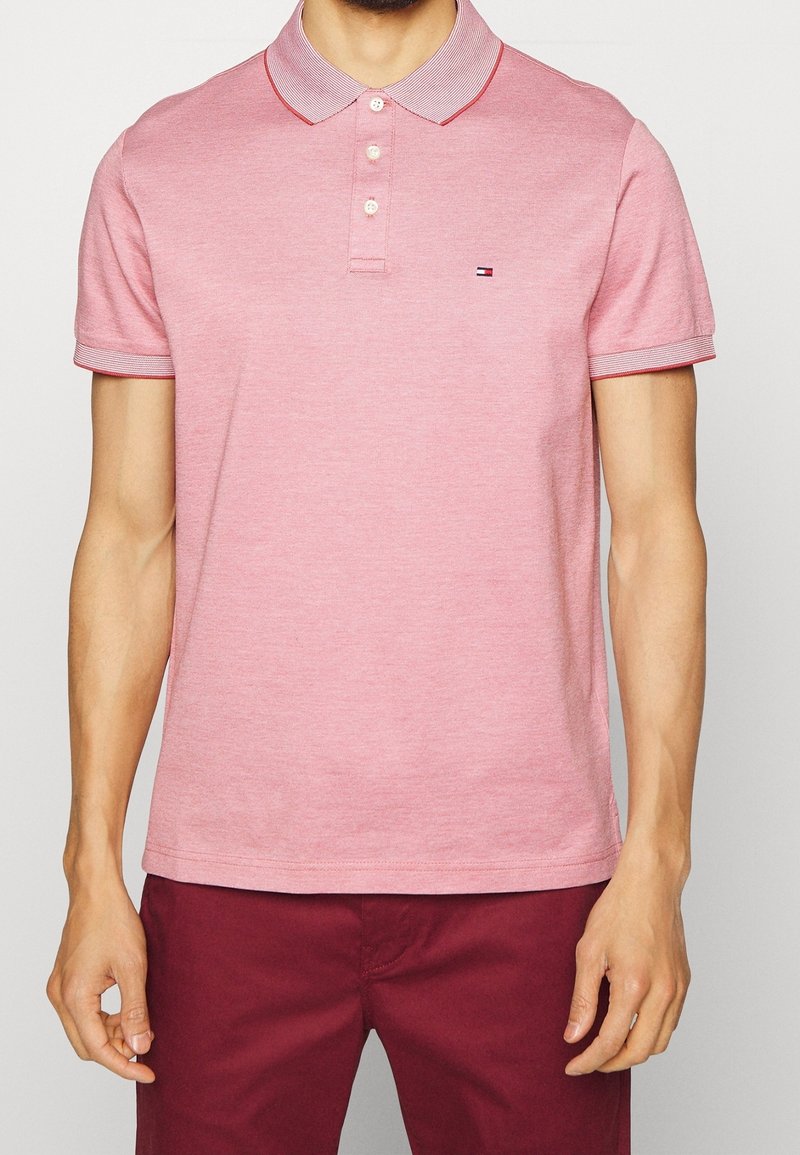 Homme portant un polo rose clair à manches courtes avec trois boutons et un petit logo, associé à un pantalon rouge foncé.
