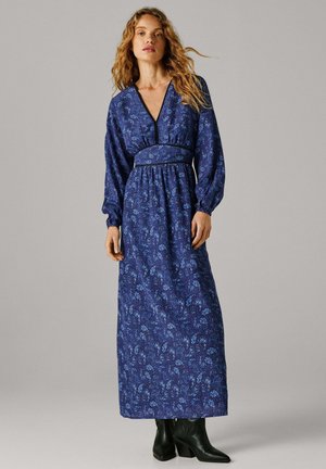 Pikk sinine kleit, millel on lillemustrid, V-kaelus ja kogutud talje. Varrukad on paugulised ning sellega kantakse musti pahkluu-saapaid.