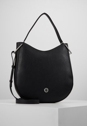 Sac à bandoulière en cuir noir avec dessus arrondi, emblème logo argenté, sangle ajustable et pièces métalliques sur une surface blanche.