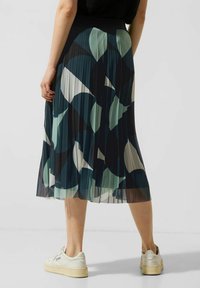 Gonna midi plissée in verde scuro e blu con motivi geometrici, dotata di una texture liscia e una vita nera. Abbinata a sneakers bianche.