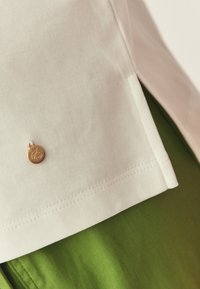 Κοντινή λήψη της ραφής λευκού υφάσματος με μικρό στρογγυλό χρυσό γούρι πάνω σε πράσινο ύφασμα.