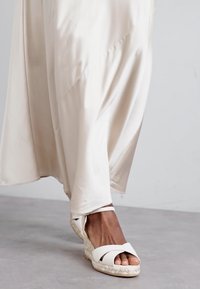 Valkoiset espadrille-korkokengät, joissa on ristikkäin sidottu muotoilu, taustalla virtaava beige mekko, jonka pinta on sileä ja satiininen.