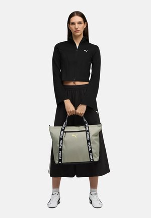 Bolso tote gris con asas negras que presenta el logo "PUMA" en blanco; modelo vistiendo una chaqueta negra corta y una falda negra, de pie.