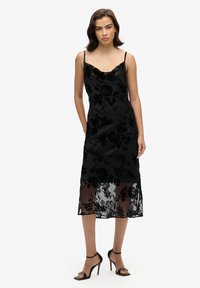 Robe en velours noir à motifs floraux avec une finition satinée, dotée d'un col drapé et de bretelles spaghetti réglables, agrémentée d'un ourlet transparent.