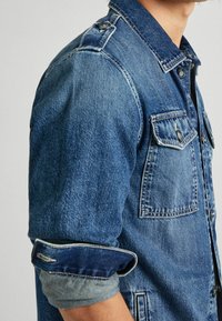 Jeansjacka i mellanmörkblå färg med klassisk krage, knappade bröstfickor och uppkavlade ärmar som avslöjar ett ljusare innerfoder.