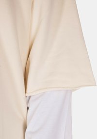 Tessuto di colore crema con una texture liscia, caratterizzato da un design semplice e una vestibilità comoda. L'orlo è rifinito con cura. Una camicia bianca è visibile sotto.