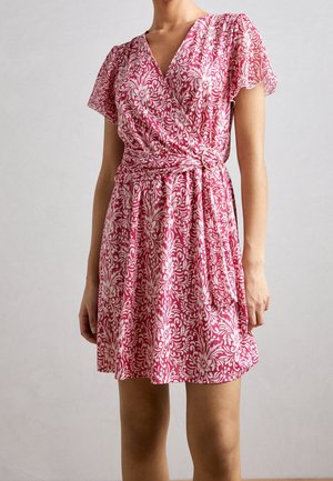 Nainen, joka käyttää polvenpituista kietaisumekkoa, jossa on lyhyet liehutetut hihat, pinkki- ja valkoinen kukkakuviointi sekä vyö, jossa on pyöreä solki vyötäröllä.