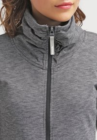 Gros plan sur une personne portant une veste zippée grise à rayures avec un col montant et un curseur de fermeture éclair portant l'inscription "Bench".