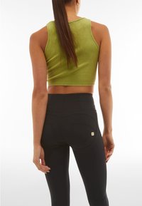 Femme aux longs cheveux portant un haut court sans manches vert et des leggings noirs taille haute, debout dos à la caméra.