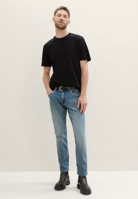 Tricou din bumbac negru, croi lejer, purtat cu blugi de denim bleu deschis. Botine negre cu curea, stând pe un fundal neutru.