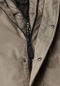 Beige jacka med svart dragkedja, silverknappar och texturerad tyg. Har vadderad foder och dragsko-detalj runt kragen.