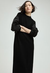 Schwarzes Langarmkleid mit figurbetontem Oberteil und voluminösen schwarzen Stoffärmeln. Glatte Textur mit minimalistischen Design und Rundhalsausschnitt.
