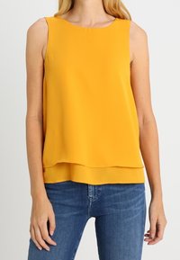 Geel mouwloos topje met een ronde halslijn, gelaagd ontwerp en lichte stof. Gecombineerd met blauwe jeans, die een eenvoudige, strakke silhouet hebben.