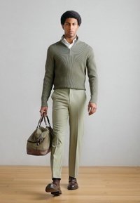 Grøn ribstrikket sweater med lynlås, lysegrønne bukser, brune sko og en brun taske med læderhåndtag. Enkel, moderne silhuet.