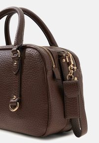 Braune strukturierte Lederhandtasche mit doppelten Griffen, goldfarbenen Reißverschlüssen und einem abnehmbaren Schulterriemenhaken vor hellem Hintergrund.