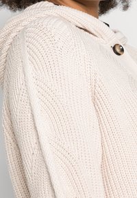 Dziana kremowa bluza z kapturem o teksturowanym wzorze, z dużym zapięciem na guzik i miękkim, zwisającym kapturem.