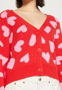 Roter Cardigan mit V-Ausschnitt und rosa Herzmustern, ausgestattet mit einem gerippten Saum und drei großen roten Knöpfen auf der Vorderseite.
