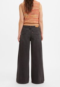 Woman wearing wide-leg black denim jeans and a sleeveless orange knit top, standing facing away against a plain white background.