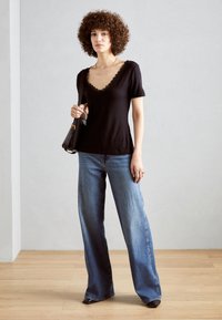 Svart kortermet topp med v-hals med blondekanter, parret med lyseblå vidt utskårne jeans. Modellen holder en svart skulderveske.