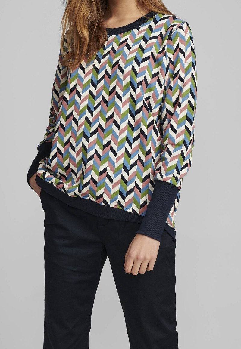 Personne portant une chemise à manches longues à motif chevron multicolore avec des poignets foncés et un pantalon foncé sur un fond gris uni.