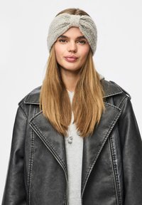 Jeune femme aux longs cheveux blonds portant un bandeau gris en tricot et une veste en cuir noire par-dessus un sweat à capuche gris clair, sur fond blanc.