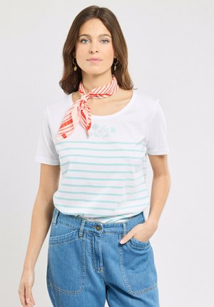 Femme aux cheveux bruns portant un t-shirt blanc à rayures, un jean bleu avec de grandes poches et un foulard rouge rayé autour du cou, debout avec une main dans la poche.