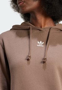 Brun hoodie med texturerat tyg, utrustad med en känguruficka, dragsko och en vit Adidas-logga på bröstet.