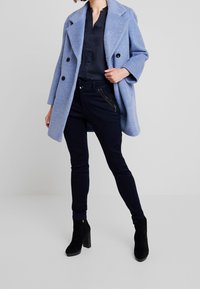 Kvinna iklädd en ljusblå ullkappa över en mörk marinblå blus och slim-fit byxor med dragkedjedetaljer, tillsammans med svarta mockaboots. 