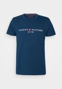 Mörkblå t-shirt med kort ärm med "Tommy Hilfiger" och logotyp i vitt och rött centrerat på bröstet, rund halsringning, storlek medium, etikett synlig inuti kragen.